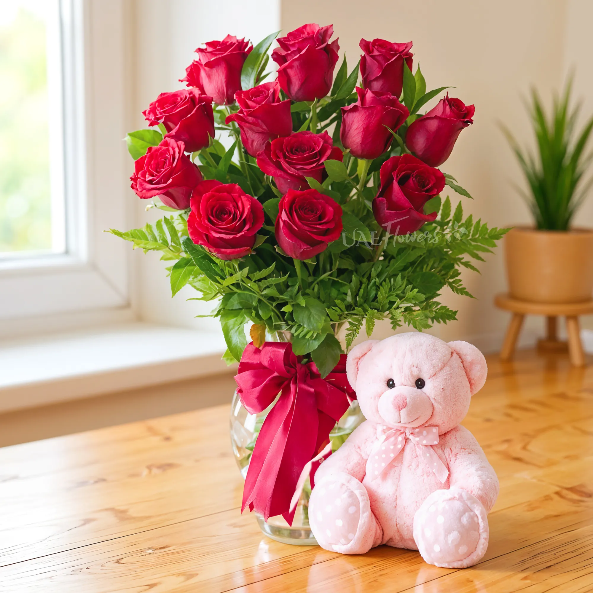 12 red roses with green fillers in a glass vase and a small pink teddy bear.
