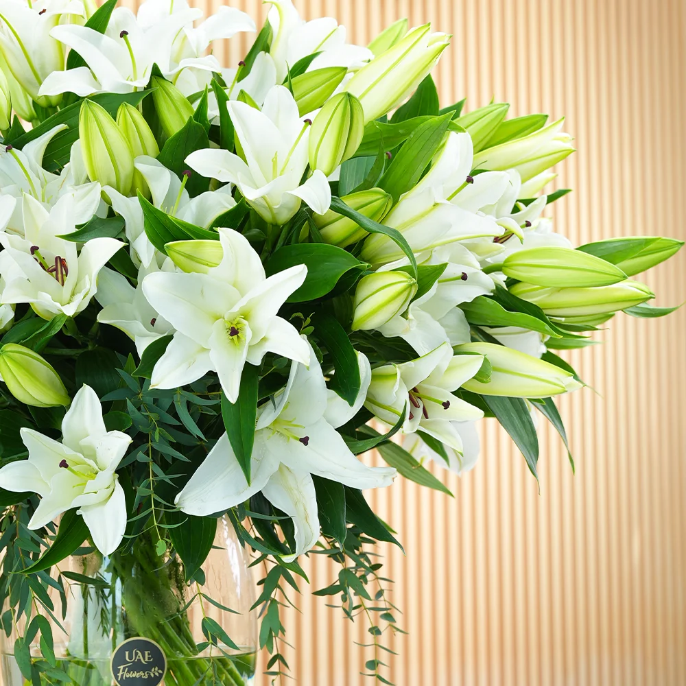 An arrangement of 20 white lilies with green fillers in a glass vase.