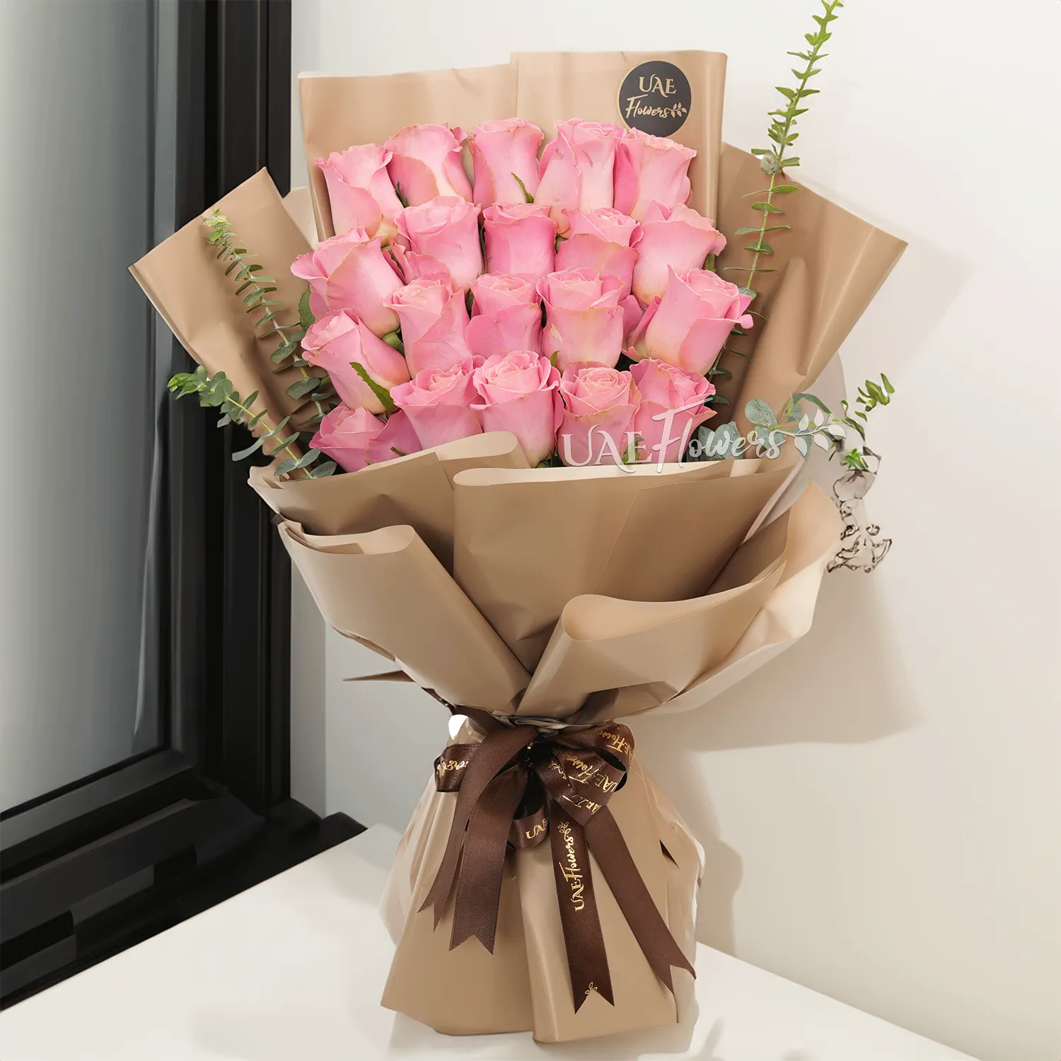 Bouquet of 20 pink roses with green fillers in paper packing tied with brown ribbons.