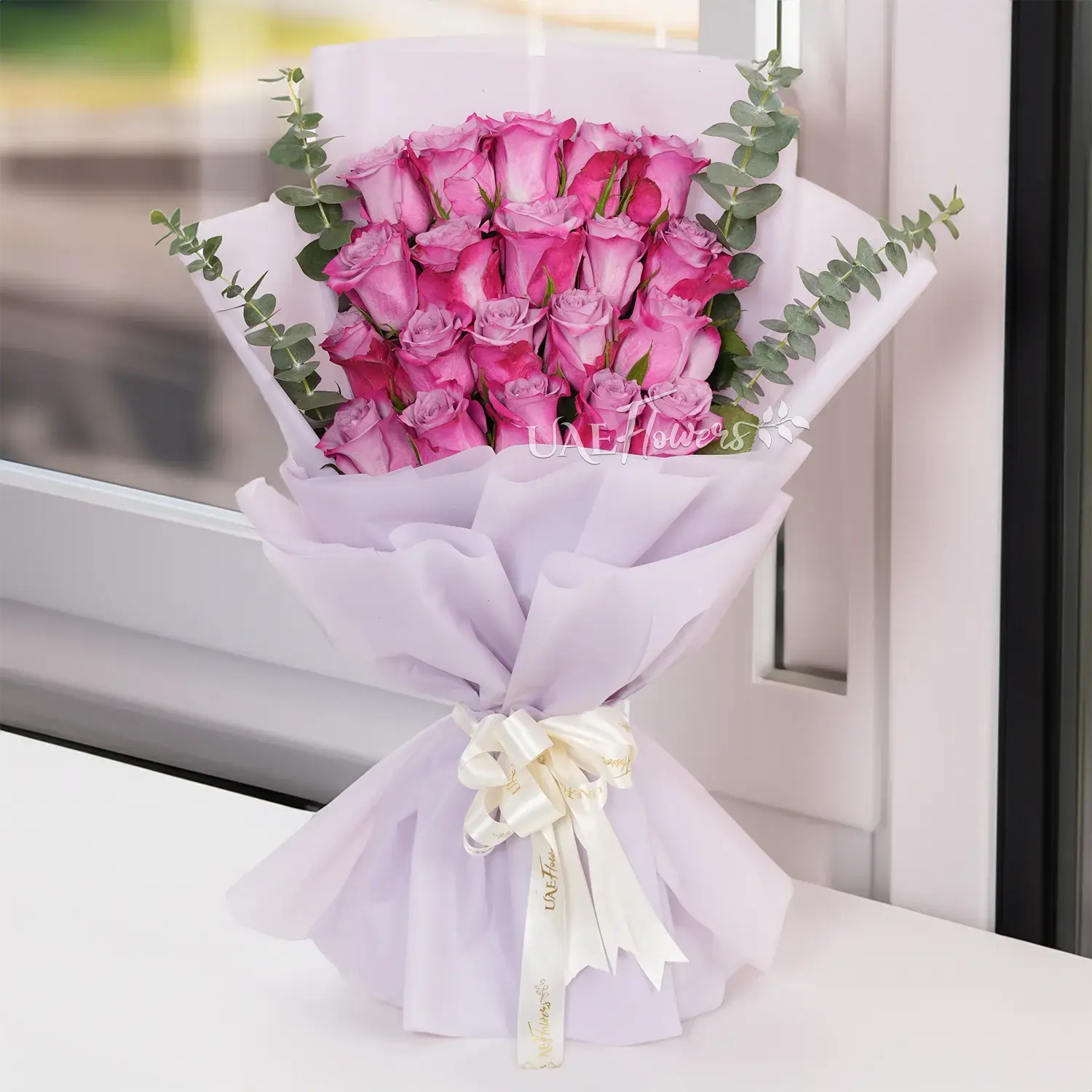 Bouquet of 20 purple roses with eucalyptus leaf in pink paper packing tied with white ribbons.