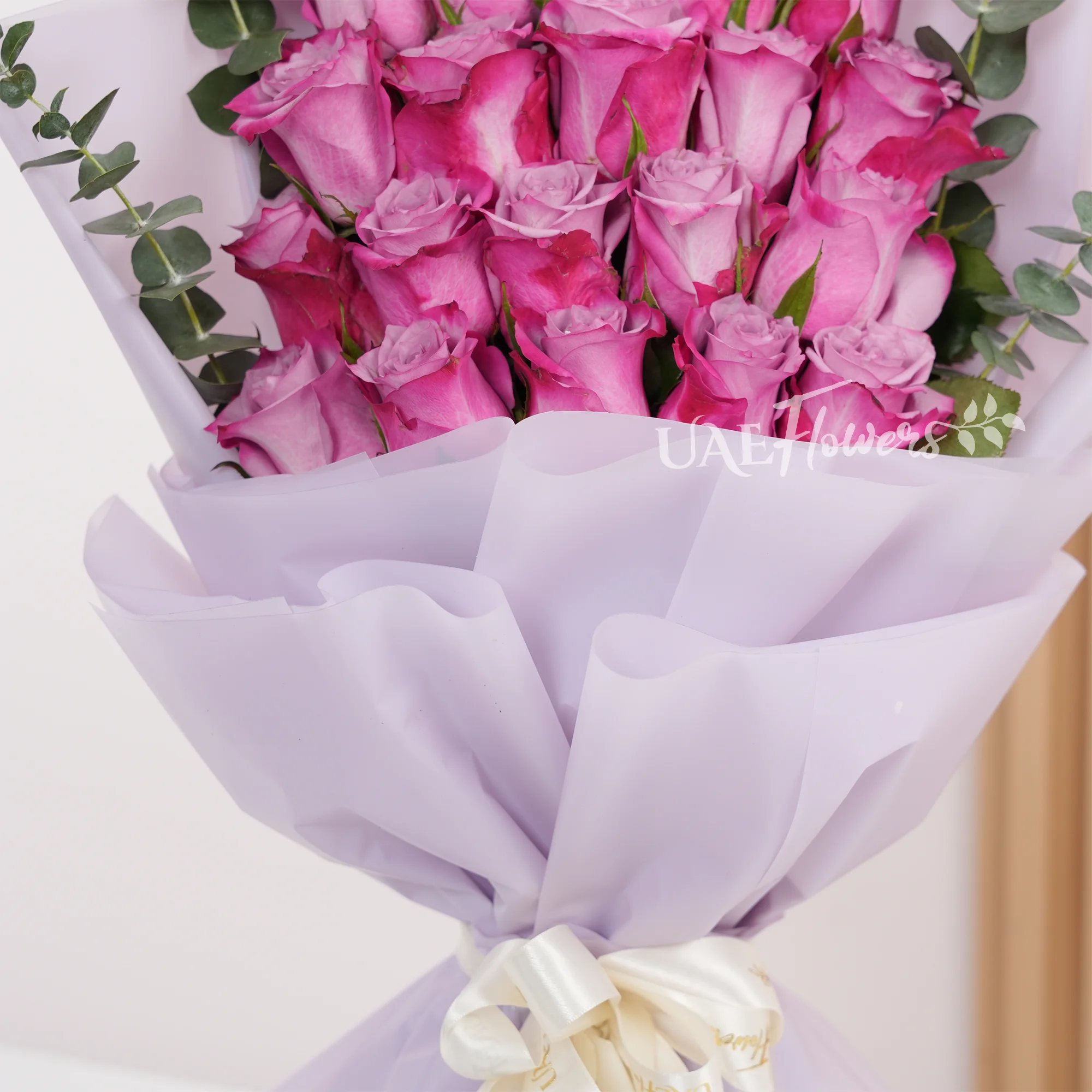 Bouquet of 20 purple roses with eucalyptus leaf in pink paper packing tied with white ribbons.