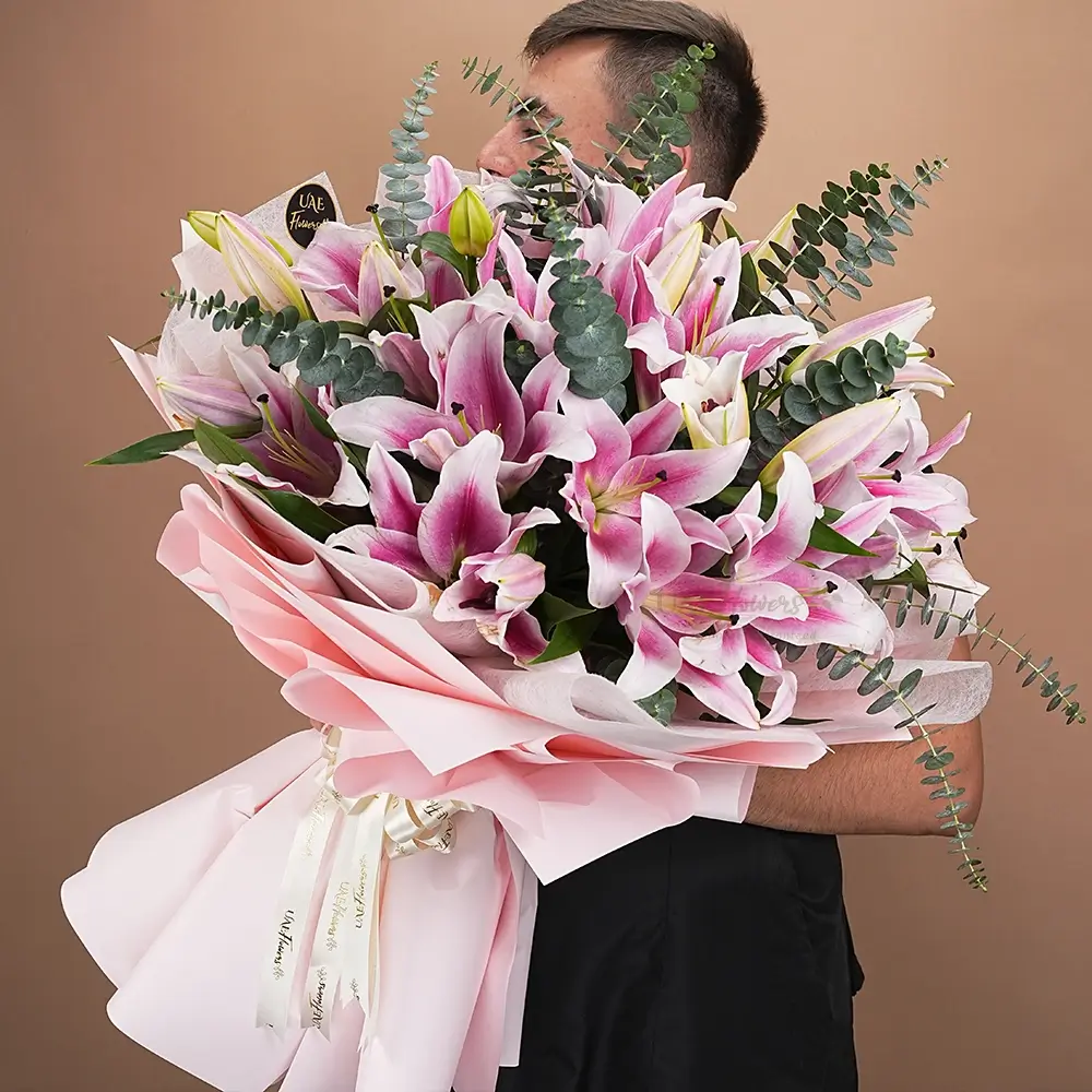 Bouquet of 10 pink lilies with eucalyptus leaf and green fillers in pink wrapping paper tied with pink ribbon.