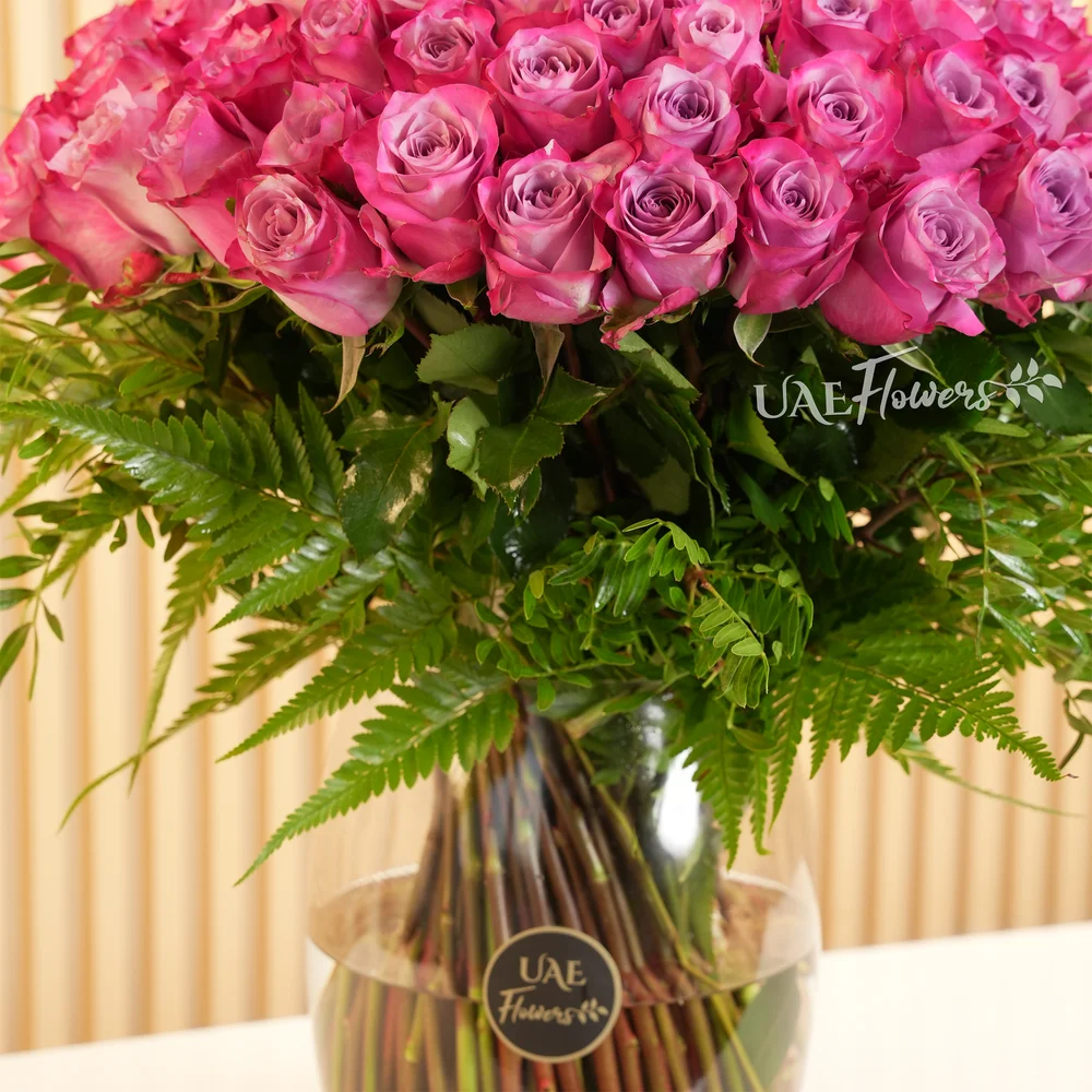 100 Purple Roses with green fillers in a glass vase.
