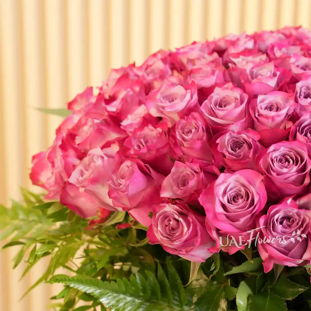 100 Purple Roses with green fillers in a glass vase.