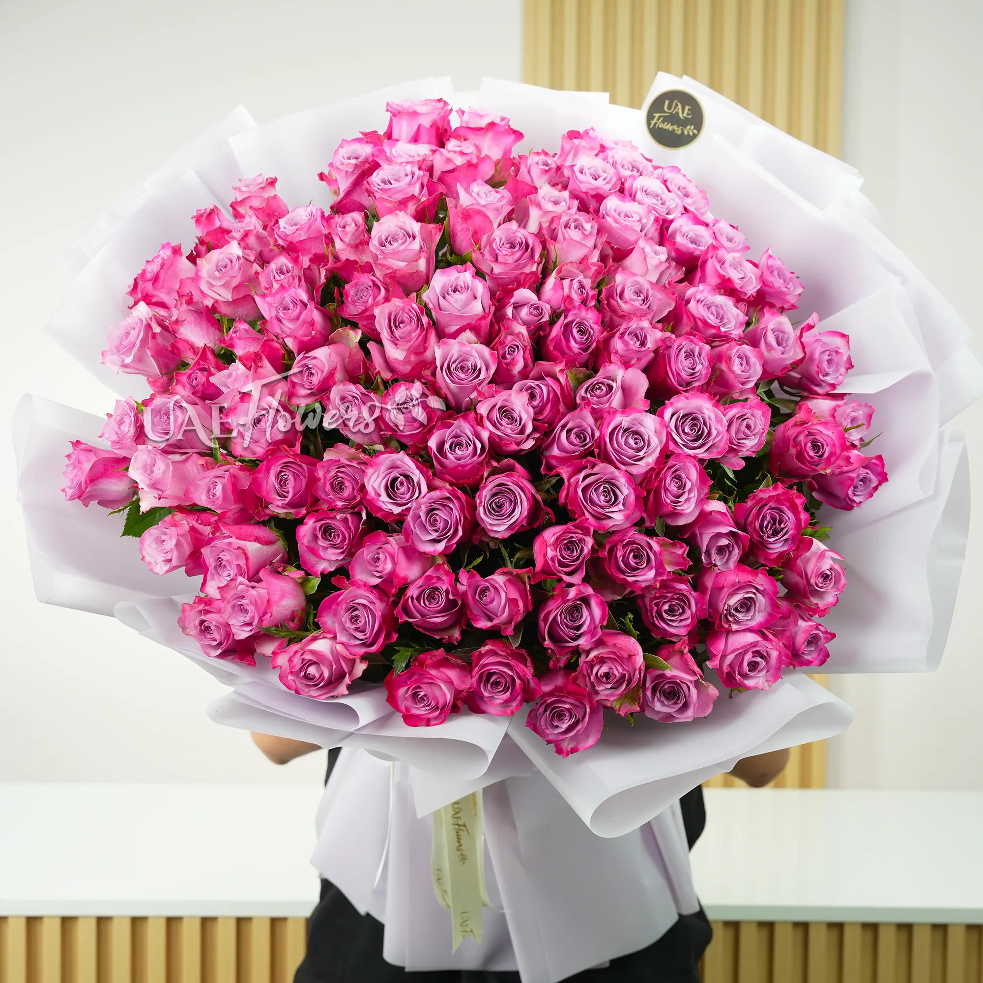 Bouquet of 100 purple roses with fillers in a white wrapping tied with ribbons.