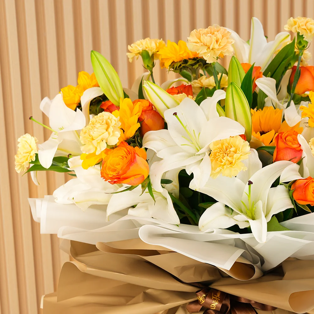 Bouquet of 5 white lilies, 5 sunflowers, 10 orange roses and 15 peach carnations with green fillers wrapped in paper packing and tied with ribbons.