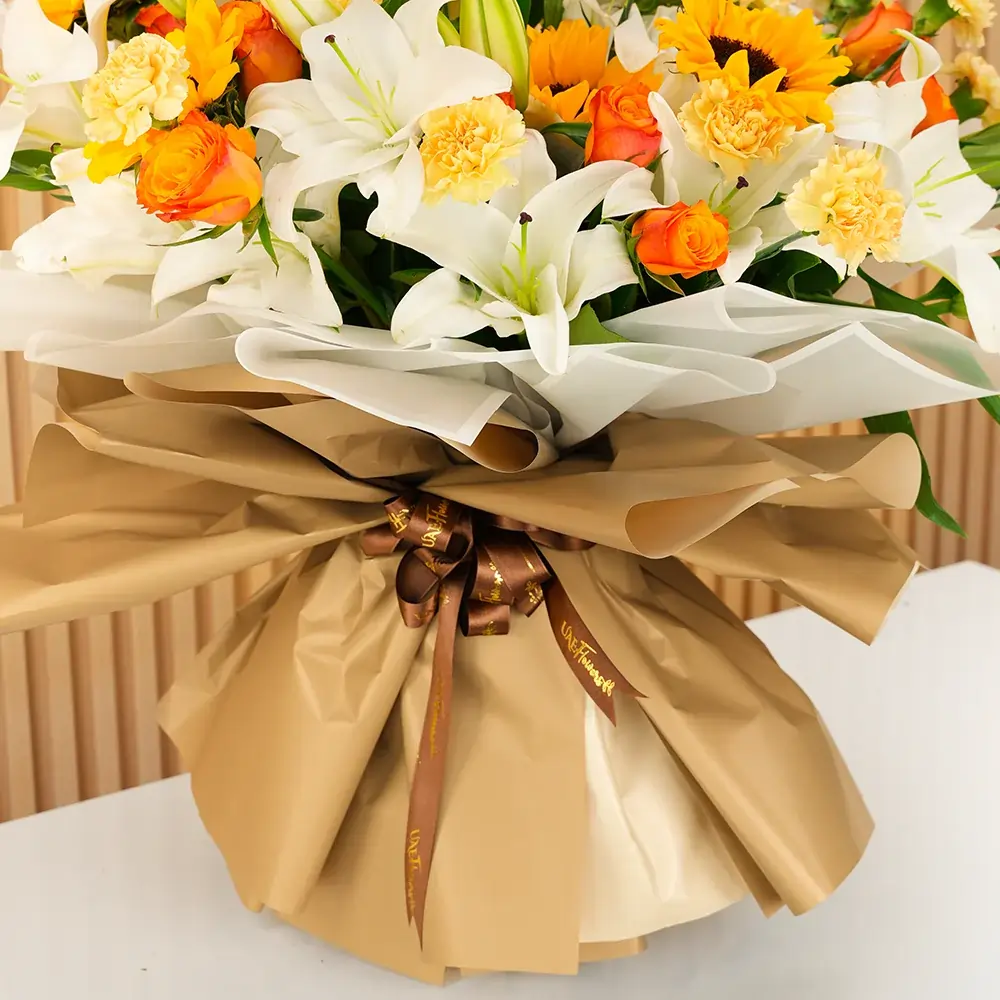 Bouquet of 5 white lilies, 5 sunflowers, 10 orange roses and 15 peach carnations with green fillers wrapped in paper packing and tied with ribbons.